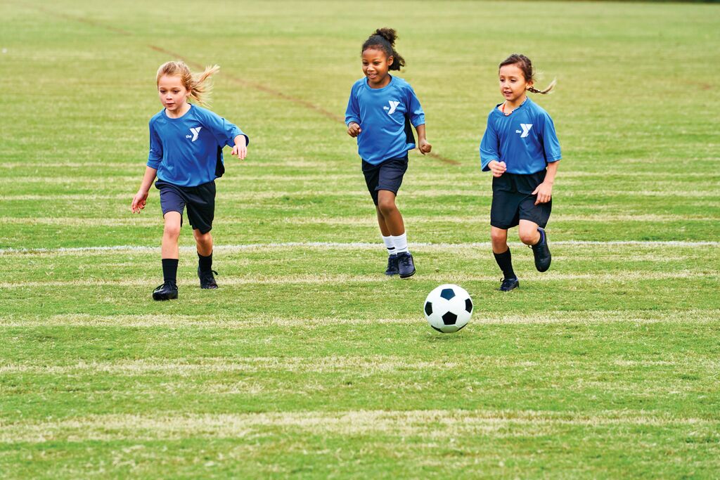 Three girls running after soccer ball on soccer field wearing YMCA logo uniforms