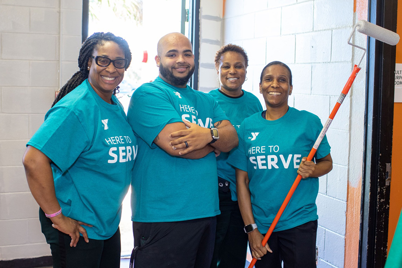 Four people in turquoise shirts featuring the YMCA logo and the words Here to Serve